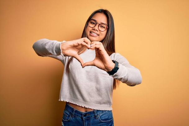 Młoda piękna azjatycka dziewczyna w luźnym swetrze i okularach na żółtym tle uśmiechając się w miłości robi kształt symbolu serca z rąk. Romantyczna koncepcja. - Zdjęcie, obraz