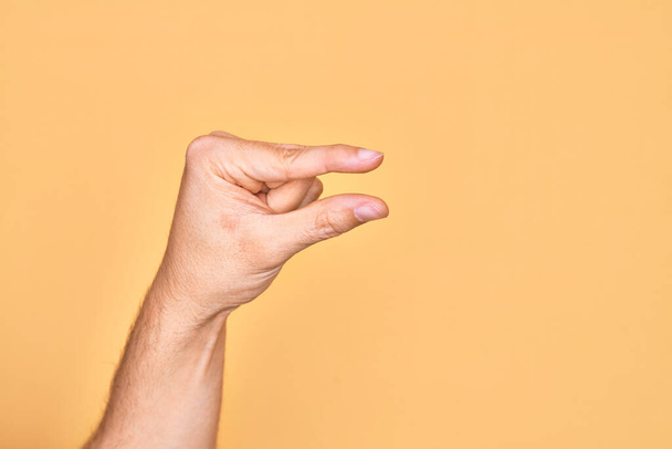 Hand of caucasian young man showing fingers over isolated yellow background picking and taking invisible thing, holding object with fingers showing space - Foto, Bild