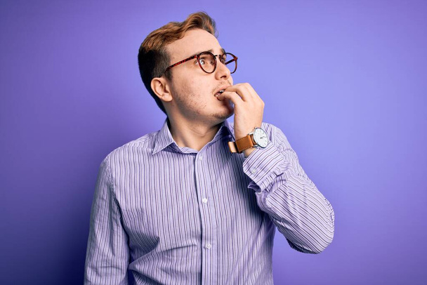 Νεαρός όμορφος κοκκινομάλλης που φοράει casual πουκάμισο και γυαλιά σε μωβ φόντο που δείχνει αγχωμένος και νευρικός με τα χέρια στο στόμα να δαγκώνουν νύχια. Πρόβλημα άγχους. - Φωτογραφία, εικόνα