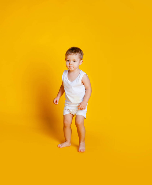 chlapec stojí v bílém tričku a kalhotkách na oranžovo-žlutém pozadí - Fotografie, Obrázek