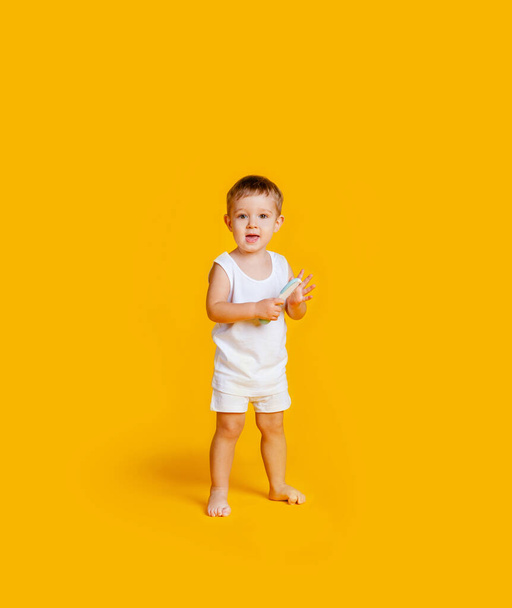 jongen staat in een wit T-shirt en onderbroek en met een kam in zijn handen op een oranje-gele achtergrond - Foto, afbeelding