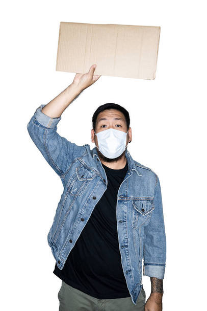Γενειοφόρος Ασιάτης εργάτης ντυμένος με τζιν με ιατρική μάσκα κρατάει χαρτόνι σε λευκό φόντο.Η έννοια της διαμαρτυρίας, της προσοχής, του αιτήματος. Τοποθετήστε για κείμενο ή χώρο αντιγραφής. Διαδρομή αποκοπής. - Φωτογραφία, εικόνα