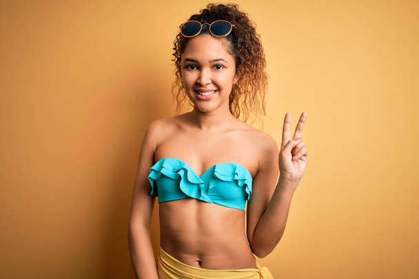 Fiatal gyönyörű amerikai nő nyaralni visel bikini felett elszigetelt sárga háttér mosolygós nézi a kamera mutató ujjak csinál győzelem jele. Második.. - Fotó, kép