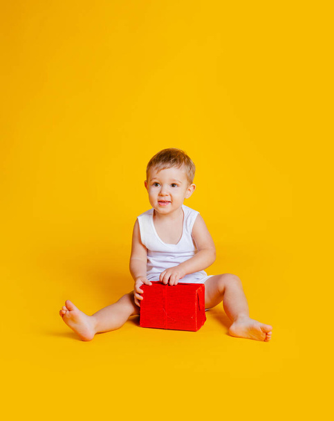 um menino senta-se em uma camiseta e cuecas ao lado de uma caixa de presente vermelha
. - Foto, Imagem