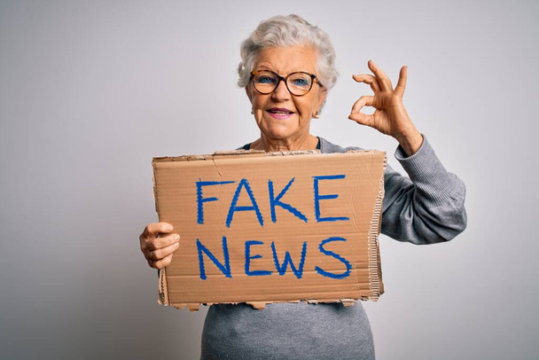 Женщина с седыми волосами, держащая баннер с фальшивыми новостями на белом фоне, делает знак ОК с пальцами, отличный символ
 - Фото, изображение