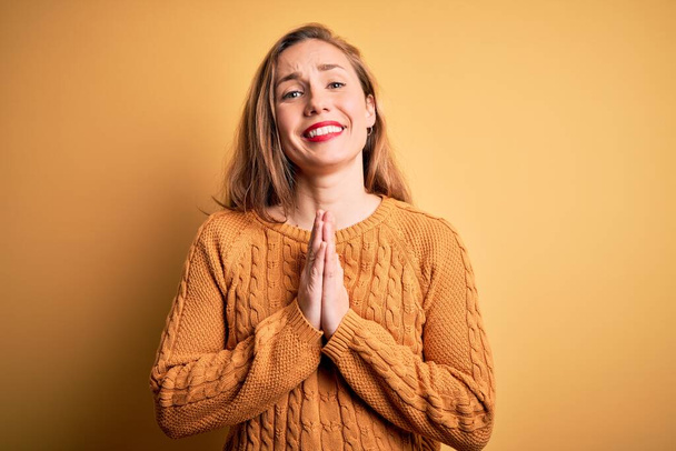 Young beautiful blonde woman wearing casual sweater standing over yellow background praying with hands together asking for forgiveness smiling confident. - Photo, Image