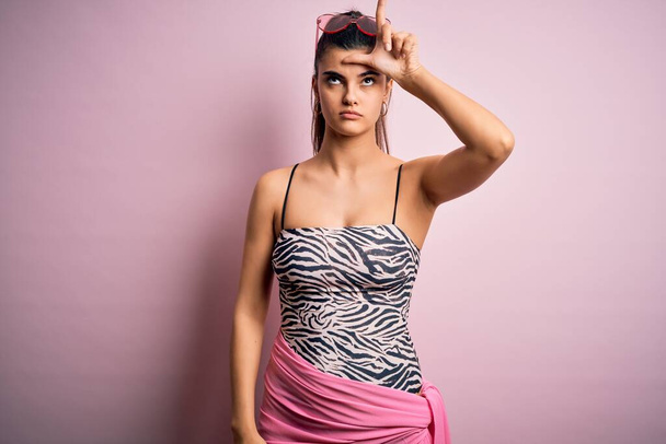 Młoda piękna brunetka na wakacjach ubrana w strój kąpielowy na różowym tle nabija się z ludzi z palcami na czole robiąc przegrany gest szydząc i obrażając. - Zdjęcie, obraz