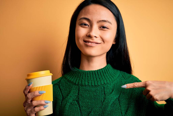 Młoda piękna Chinka pije kawę na odizolowanym żółtym tle z zaskakującą twarzą wskazującą palcem na siebie - Zdjęcie, obraz