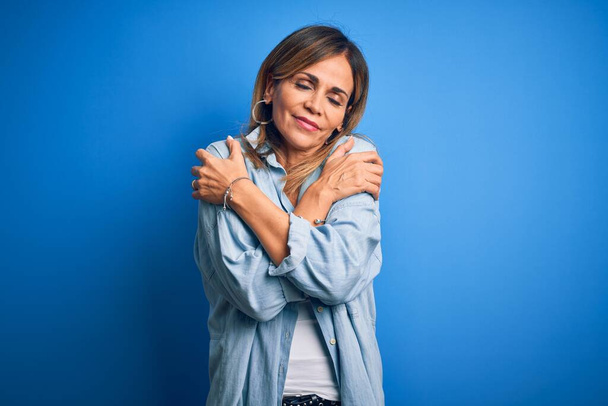 Middelbare leeftijd mooie vrouw dragen casual shirt staan over geïsoleerde blauwe achtergrond knuffelen zichzelf gelukkig en positief, glimlachen zelfverzekerd. Zelfliefde en zelfzorg - Foto, afbeelding