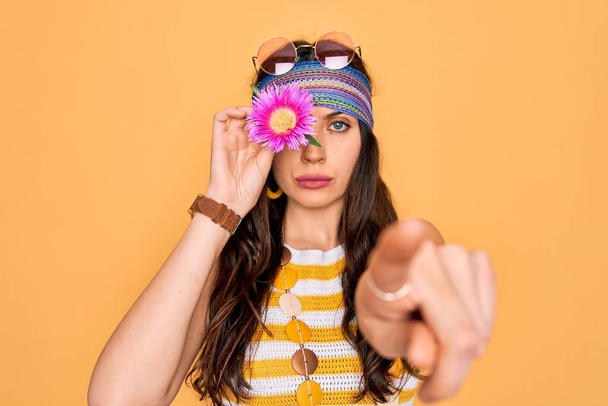 Joven mujer hippie hermosa con ojos azules con gafas de sol sosteniendo flor rosa sobre el ojo señalando con el dedo a la cámara y a usted, signo de mano, gesto positivo y seguro de la parte delantera
 - Foto, Imagen