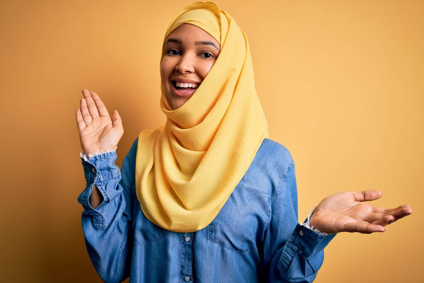 Junge schöne Frau mit lockigem Haar, die einen traditionellen arabischen Hijab über gelbem Hintergrund trägt und fröhlich lächelnd Hände anbietet, die Hilfe und Akzeptanz geben. - Foto, Bild