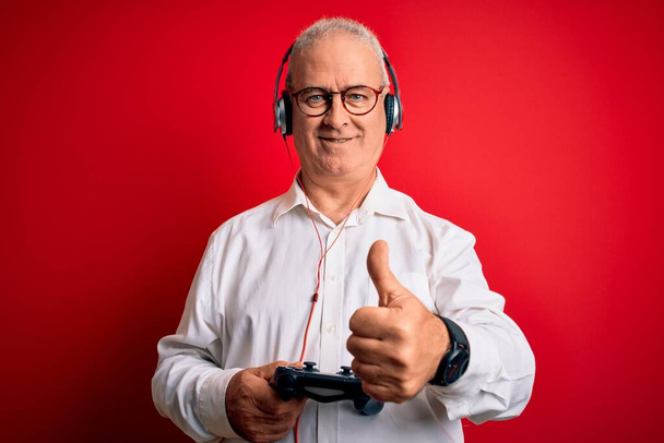 Красивий чоловік середнього віку, який грає у відеоігри, використовує джойстик та навушники, задоволені великою посмішкою, роблячи знак ок, великий палець пальцями, відмінний знак
 - Фото, зображення