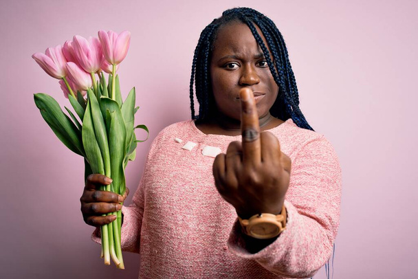 Joven afroamericano más tamaño mujer con trenzas celebración ramo de tulipanes rosados flor Mostrando dedo medio, descortés y grosero joder expresión
 - Foto, imagen