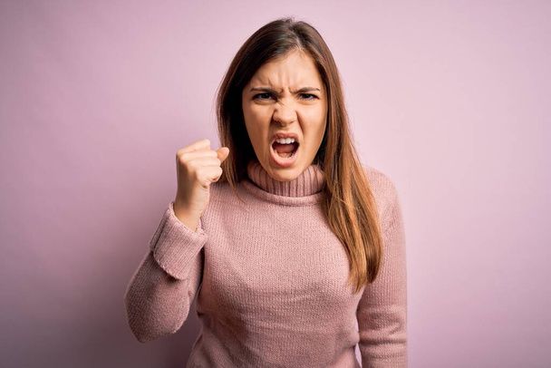 Beautiful young woman wearing turtleneck sweater over pink isolated background angry and mad raising fist frustrated and furious while shouting with anger. Rage and aggressive concept. - Foto, imagen