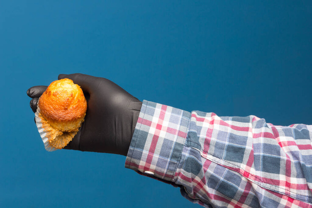Cupcake industriale in mano disinfettato e protetto con un guanto nero, su sfondo blu. Pasticceria industriale
 - Foto, immagini