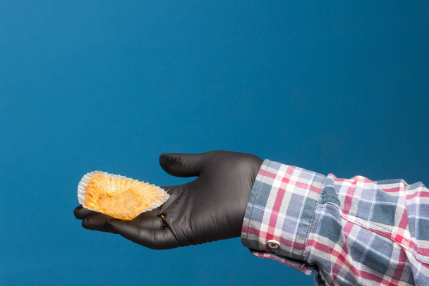 Industrieller Cupcake in der Hand desinfiziert und mit einem schwarzen Handschuh geschützt, auf blauem Hintergrund. Industriegebäck - Foto, Bild