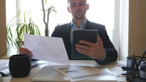 Successful businessman looking at diagrams with statistics on papers while working on digital device. Young entrepreneur analyzing statistical data information on a tablet pc in office. Dolly shot - Video