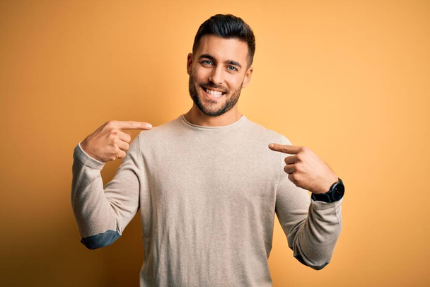 Молодой красивый мужчина в случайном свитере, стоящий на изолированном желтом фоне, уверенно выглядящий с улыбкой на лице, указывающий на себя пальцами гордым и счастливым
. - Фото, изображение