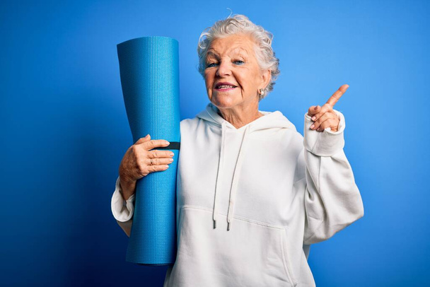 Senior schöne sportliche Frau hält Matte für Yoga steht über isolierten blauen Hintergrund überrascht mit einer Idee oder Frage Zeigefinger mit glücklichem Gesicht, Nummer eins - Foto, Bild