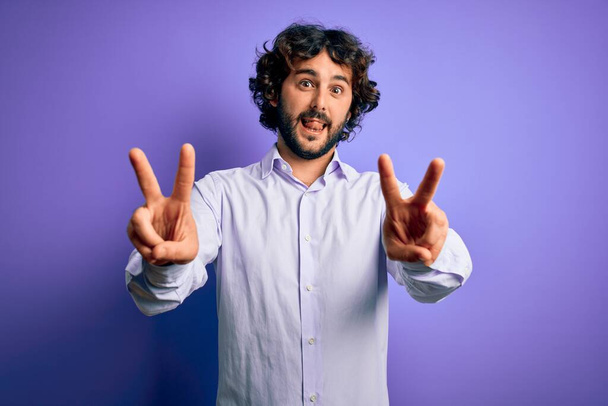 Młody przystojny biznesmen z brodą w koszuli stojący nad fioletowym tłem uśmiechnięty językiem pokazujący palce obu rąk robiących znak zwycięstwa. Numer dwa.. - Zdjęcie, obraz