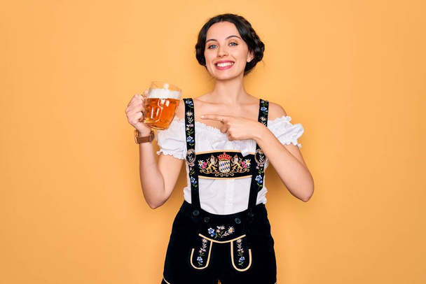 Bella donna tedesca con gli occhi azzurri che indossa il tradizionale vestito octoberfest bere vaso di birra molto felice che punta con mano e dito
 - Foto, immagini