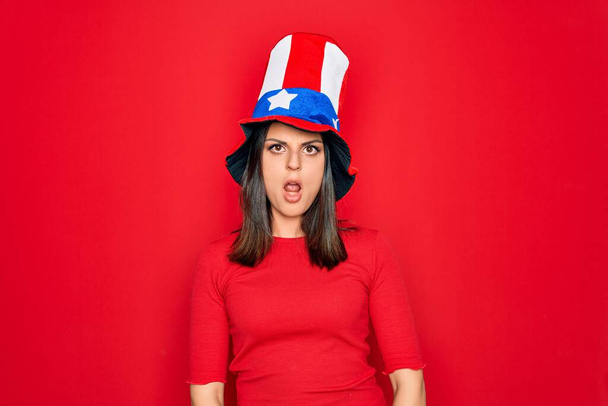 Jeune belle femme brune portant un chapeau des États-Unis célébrant la journée de l'indépendance Dans le visage choqué, l'air sceptique et sarcastique, surpris avec la bouche ouverte
 - Photo, image