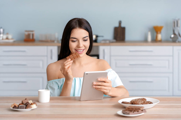 Beautiful young woman with tablet computer eating chocolate in kitchen - Photo, Image