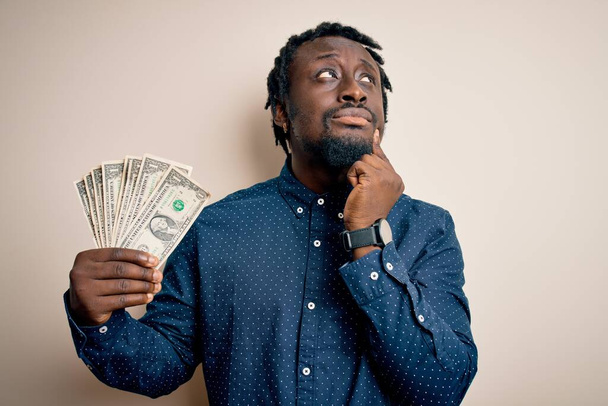 Молодий афроамериканець, який тримає купку банкнот в доларах над ізольованим білим фоном, серйозно думає про питання, дуже заплутана ідея. - Фото, зображення