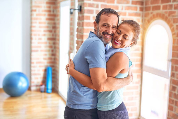 Edad media hermosa pareja deportiva sonriendo feliz. De pie con sonrisa en la cara abrazándose en el gimnasio
 - Foto, Imagen