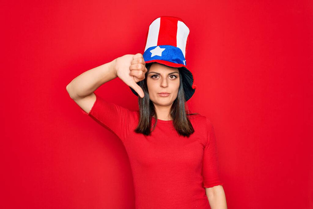 Nuori kaunis brunette nainen yllään Yhdysvalloissa hattu juhlii itsenäisyyspäivä etsivät onneton ja vihainen osoittaa hylkääminen ja negatiivinen peukalot alas ele. Huono ilme
. - Valokuva, kuva