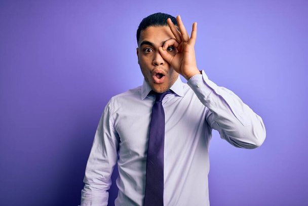 Молодой бразильский бизнесмен, одетый в элегантный галстук, стоящий на изолированном фиолетовом фоне, делает нормальный жест, шокированный удивленным лицом, глазами, смотрящими сквозь пальцы. Неверное выражение
. - Фото, изображение