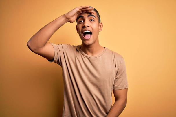 Young handsome african american man wearing casual t-shirt standing over yellow background very happy and smiling looking far away with hand over head. Searching concept. - Photo, Image
