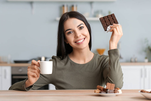 Beautiful young woman eating chocolate in kitchen - Zdjęcie, obraz