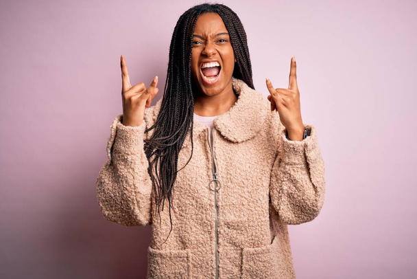 ピンクの隔離された背景の叫びにファッション冬のコートを身に着けている若いアフリカ系アメリカ人女性は手を上げて岩のシンボルをやって狂気の表情で叫びます。音楽スター。重い概念. - 写真・画像