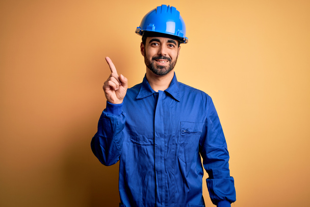 Mechaniker mit Bart, blauer Uniform und Schutzhelm über gelbem Hintergrund, der mit erhobenem Zeigefinger auf eine erfolgreiche Idee hinweist. Aufgeregt und glücklich. Nummer eins. - Foto, Bild