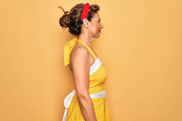 Старшие пин-ап женщина среднего возраста носить ретро-платье стиле 50-х годов на желтом фоне глядя в сторону, расслабиться профиль позировать с естественным лицом и уверенной улыбкой
. - Фото, изображение