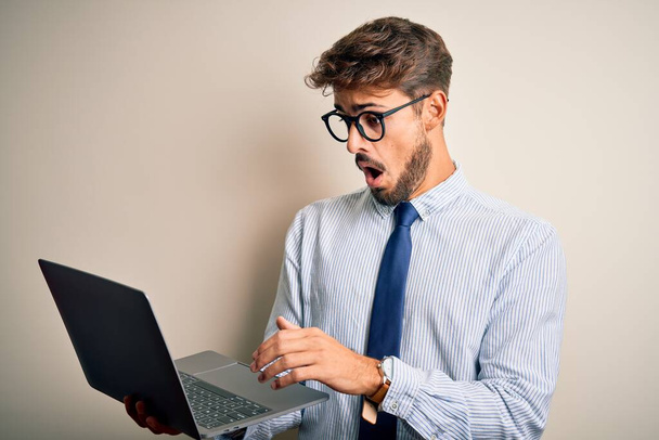 Young businessman wearing glasses working using laptop standing over white background scared in shock with a surprise face, afraid and excited with fear expression - Fotó, kép