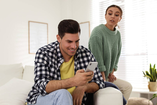 Distrustful woman peering into boyfriend's smartphone at home. Jealousy in relationship - Фото, изображение