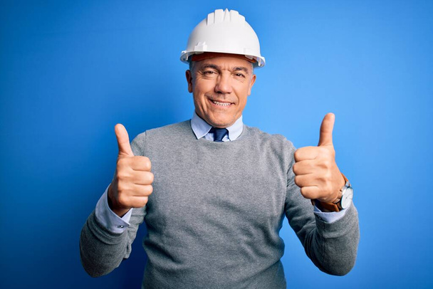 Middle age handsome grey-haired engineer man wearing safety helmet over blue background success sign doing positive gesture with hand, thumbs up smiling and happy. Cheerful expression and winner gesture. - Foto, imagen