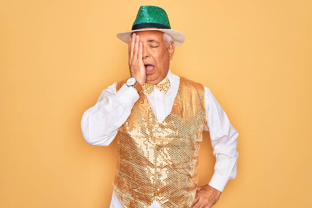 Älterer, grauhaariger Mann mittleren Alters mit brasilianischem Karnevalsgewand vor gelbem Hintergrund, gähnend müde, der das halbe Gesicht, Augen und Mund mit der Hand bedeckt. Gesicht schmerzt vor Schmerzen. - Foto, Bild