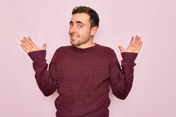 Young handsome man with blue eyes wearing casual sweater standing over pink background smiling showing both hands open palms, presenting and advertising comparison and balance - Fotografie, Obrázek