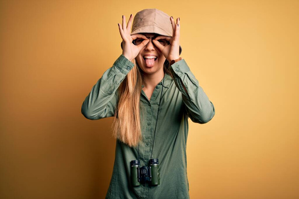 Hermosa rubia explorador mujer con ojos azules usando sombrero y gafas usando binoculares haciendo buen gesto como los binoculares pegando la lengua, los ojos mirando a través de los dedos. Exclamación absurda. - Foto, imagen