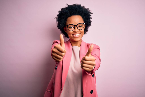 Mladá krásná afro americká byznysmena s kudrnatými vlasy v růžové bundě, která schvaluje pozitivní gesto rukou, palce nahoru, usmívá se a je šťastná za úspěch. Gesto vítěze. - Fotografie, Obrázek