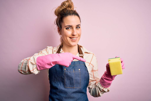 Mooie blonde schonere vrouw doet huishoudelijk werk dragen arpon en handschoenen met behulp van geurmaker zeer gelukkig wijzend met de hand en vinger - Foto, afbeelding