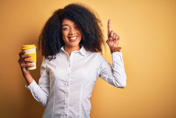 Jovem mulher de negócios afro-americana com cabelo afro bebendo café de tirar copo surpreso com uma ideia ou pergunta apontando dedo com cara feliz, número um
 - Foto, Imagem