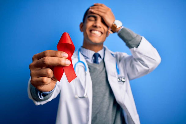 Young handsome african american doctor man holding red HIV ribbon symbol stressed with hand on head, shocked with shame and surprise face, angry and frustrated. Fear and upset for mistake. - Fotografie, Obrázek