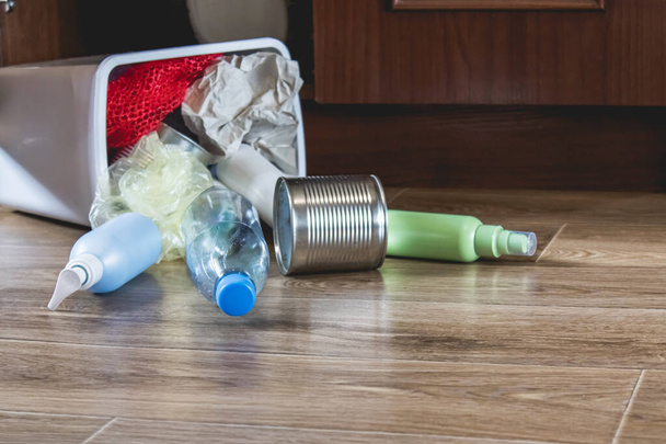 A plastic trash can with garbage lies on the floor near the kitchen cabinet - bottles, plastic and metal cans scattered in the kitchen. Garbage waste. - Fotoğraf, Görsel