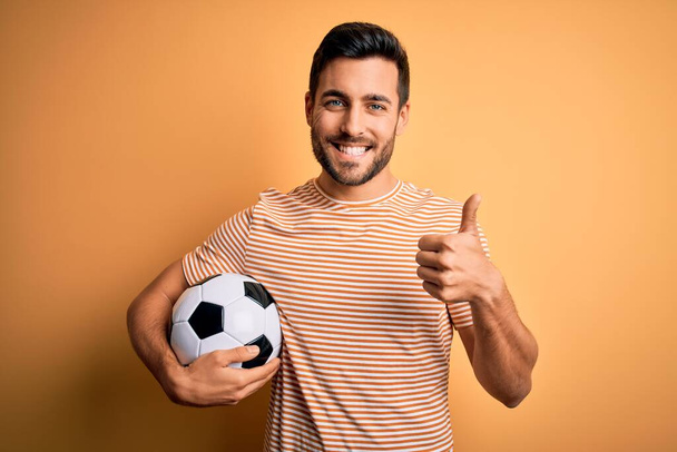 Όμορφος παίκτης με γενειάδα παίζει ποδόσφαιρο κρατώντας μπάλα ποδοσφαίρου πάνω από κίτρινο φόντο κάνει ευτυχισμένη αντίχειρες επάνω χειρονομία με το χέρι. Έγκριση έκφρασης κοιτάζοντας την κάμερα που δείχνει επιτυχία. - Φωτογραφία, εικόνα