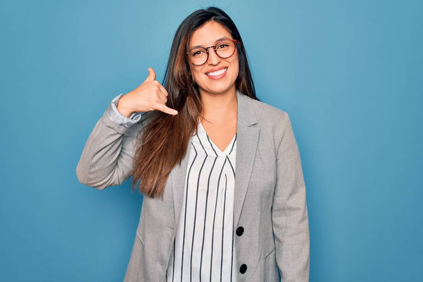 Joven mujer de negocios hispana usando anteojos parados sobre fondo azul aislado sonriendo haciendo gesto telefónico con la mano y los dedos como hablando por teléfono. Comunicar conceptos
. - Foto, Imagen