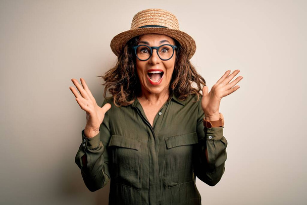 Middle age brunette woman wearing glasses and hat standing over isolated white background celebrating crazy and amazed for success with arms raised and open eyes screaming excited. Winner concept - Fotoğraf, Görsel